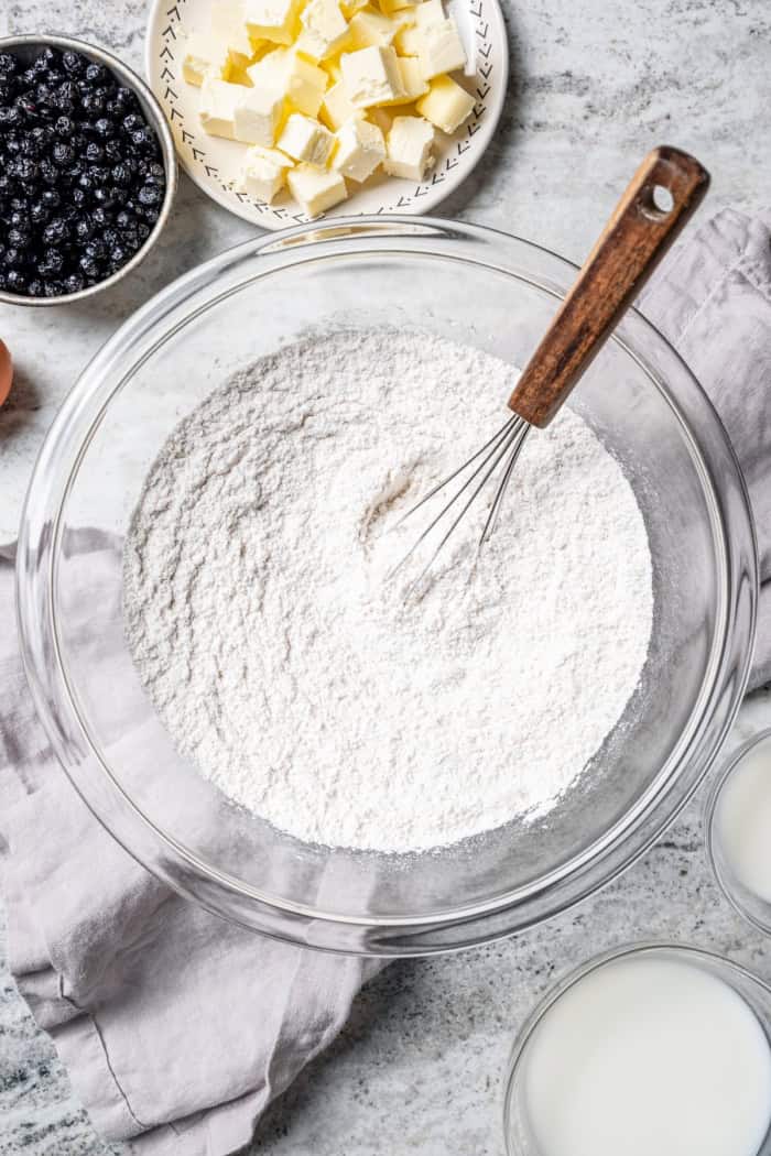 Rich, flaky and dotted with chewy dried blueberries, these blueberry scones are a bakery-style treat you can easily make at home! Overhead view of dry ingredients in mixing bowl with whisk, bowl of butter, bowl of dried blueberries, and bowl of buttermilk