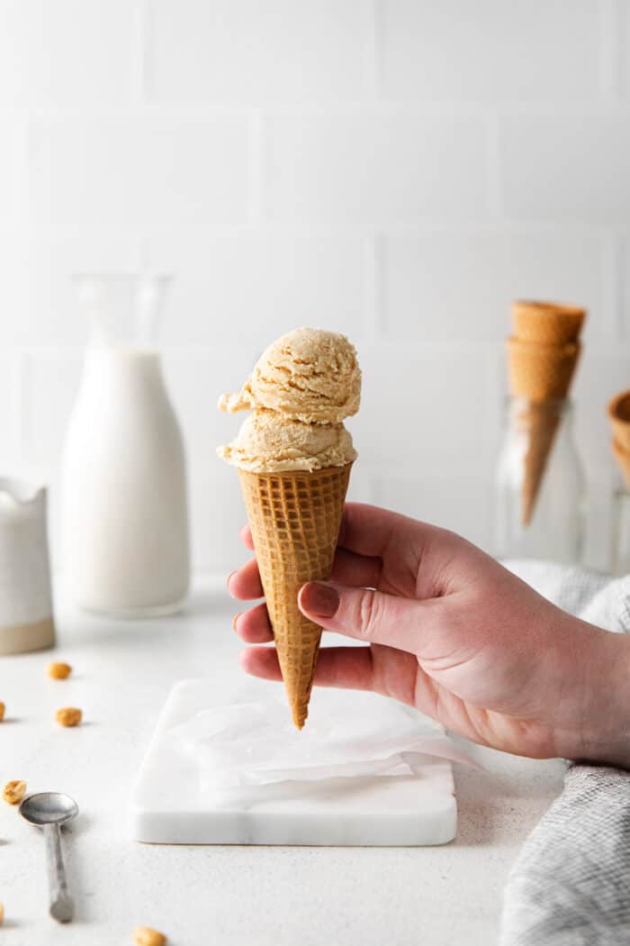 Made with just 5 ingredients and an ice cream maker, this Homemade Peanut Butter Ice Cream is sweet, creamy, and easy to make. A hand holding a waffle cone with scoops of homemade peanut butter ice cream