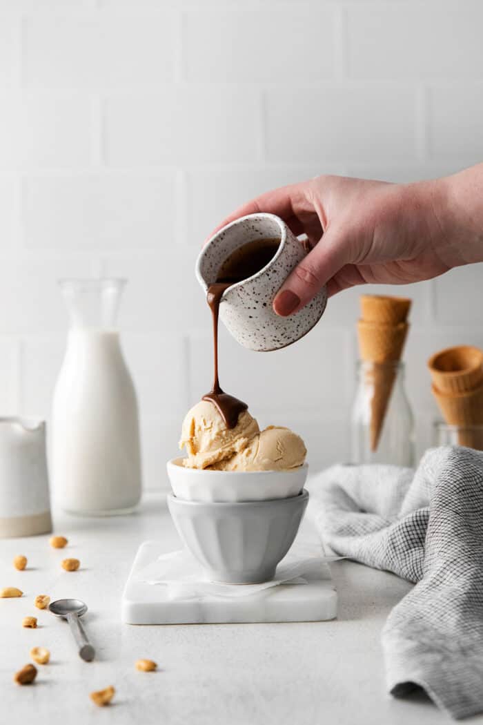 Made with just 5 ingredients and an ice cream maker, this Homemade Peanut Butter Ice Cream is sweet, creamy, and easy to make. A hand pouring hot fudge sauce over peanut butter ice cream