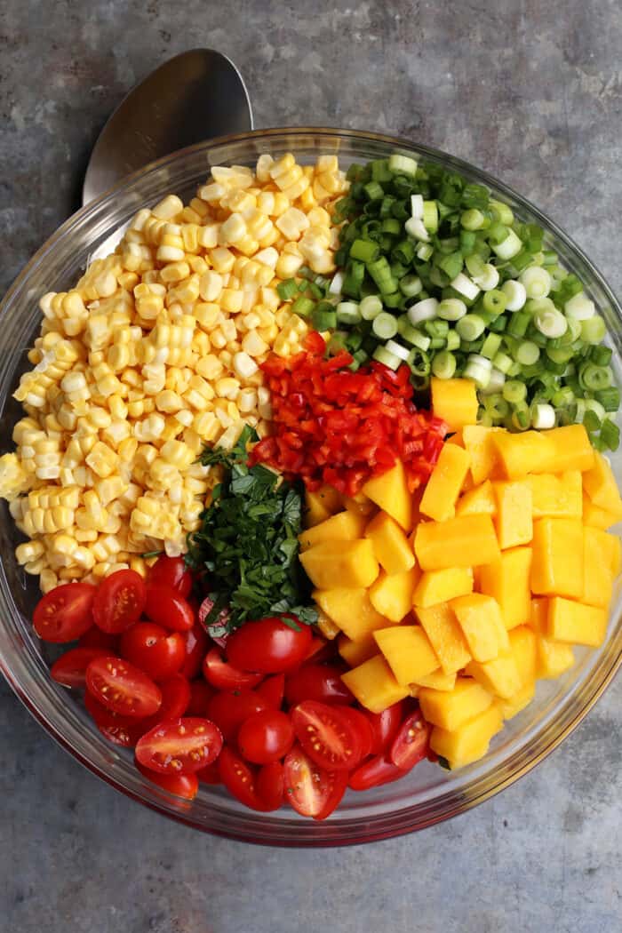 With fresh corn, mangos, tomatoes, and a touch of heat, plus a light and flavorful dressing, this Sweet Corn Salad is a summer treat! All the ingredients of sweet corn salad in a glass bowl