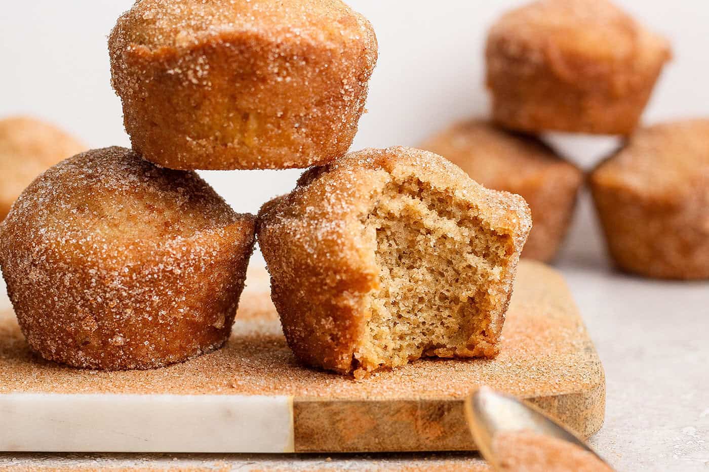 Three cinnamon sugar doughnut muffins on wooden board, with one bitten into to show moist crumb