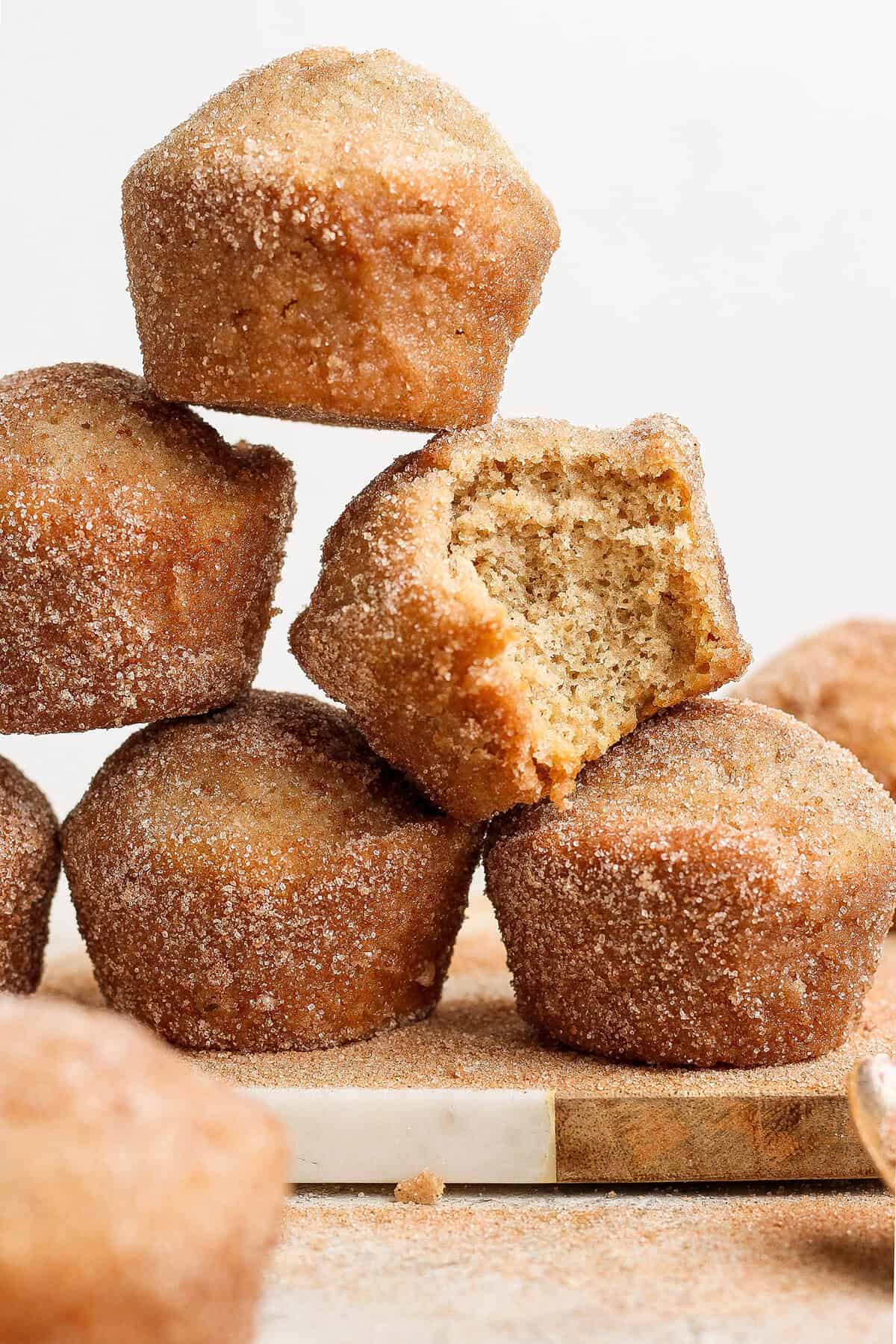 Stack of cinnamon sugar doughnut muffins on wood board, with one muffin bitten into to show moist interior
