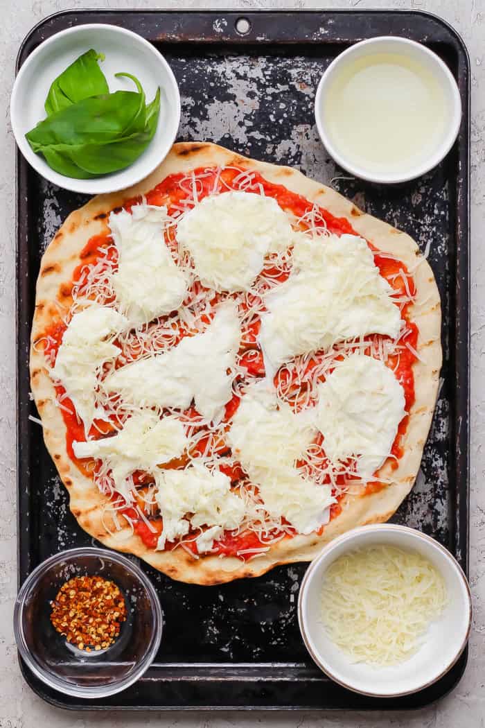 Overhead view of grilled margherita pizza on sheet pan with bowls of toppings.
