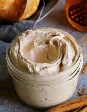 A jar of whipped cinnamon honey butter with a basket of popovers and a jar of honey in the background.