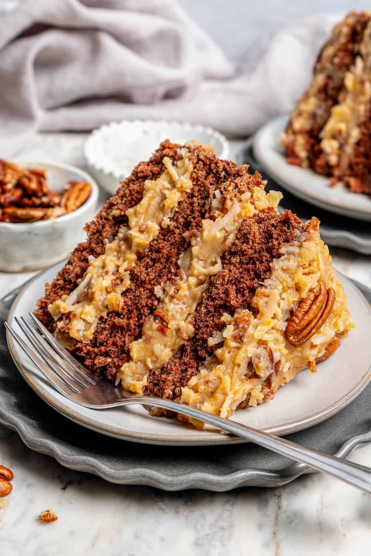 This classic German Chocolate Cake recipe is a moist, rich three-layer chocolate cake topped with a nutty coconut-pecan frosting. Perfection! Slice of classic German chocolate cake on plate with fork