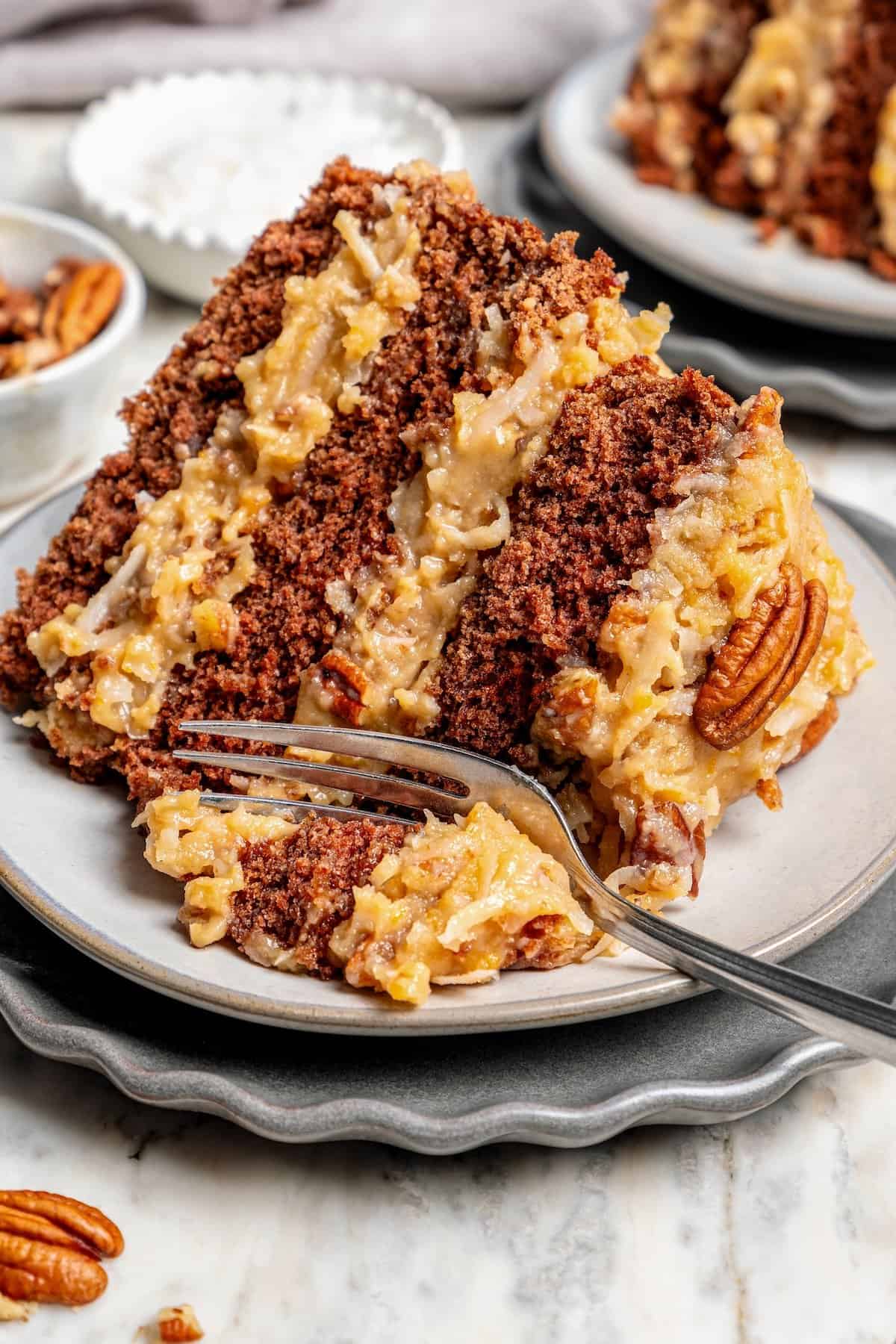 This classic German Chocolate Cake recipe is a moist, rich three-layer chocolate cake topped with a nutty coconut-pecan frosting. Perfection! Fork digging into slice of German chocolate cake