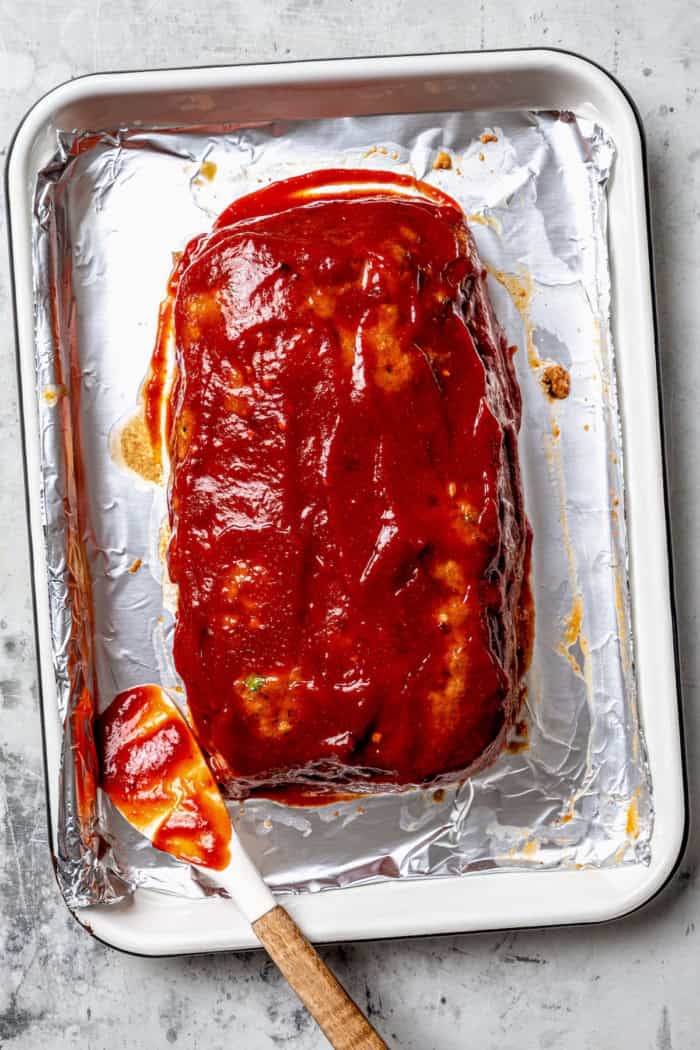 Spicy Meatloaf is a classic with a kick! It has a little Tex-Mex flair thanks to cumin and jalapeño, with a chili glaze to bring the heat. Spicy meatloaf sauce spread onto meatloaf on pan.
