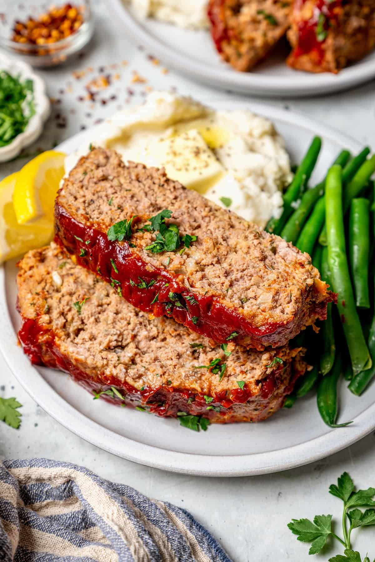 Spicy Meatloaf is a classic with a kick! It has a little Tex-Mex flair thanks to cumin and jalapeño, with a chili glaze to bring the heat. Two slices of spicy meatloaf on plate with mashed potatoes and green beans.