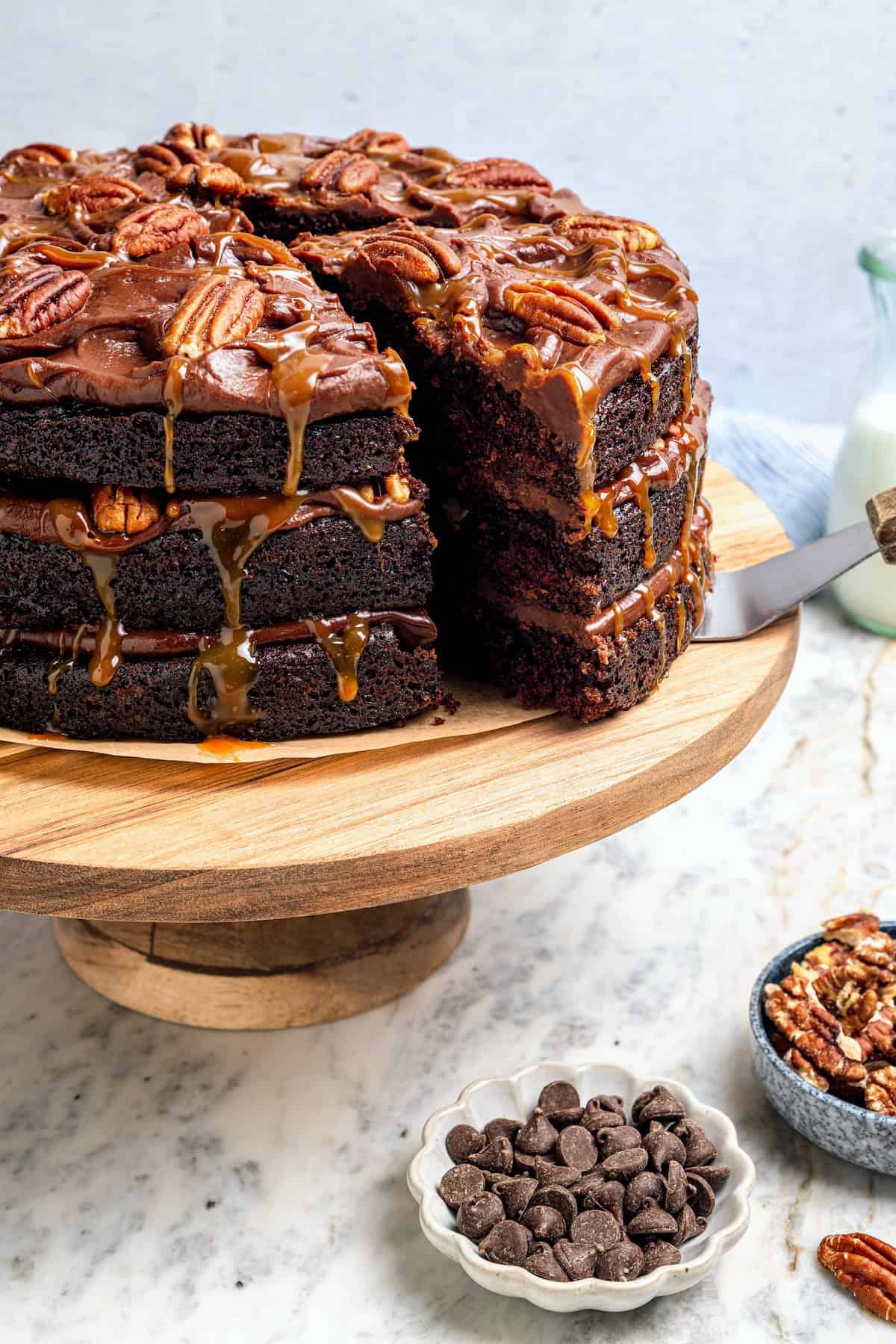 Three layers of dark chocolate cake are smothered with rich fudgy frosting, creamy caramel, and crispy salted pecans. So good! Pulling slice of turtle cake from cake stand.