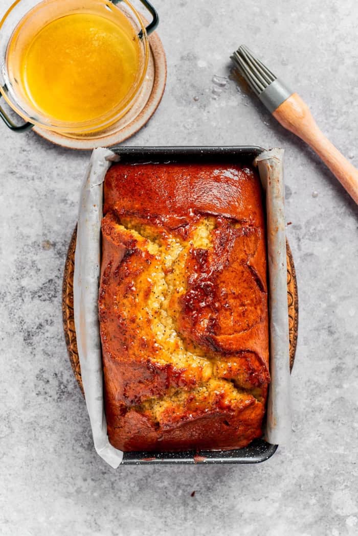 This moist orange poppy seed bread is soaked in a zippy citrus glaze for a bright, tender, bakery-style loaf perfect for brunch or gifting. Loaf of orange poppy seed bread in pan with bowl of glaze and brush.