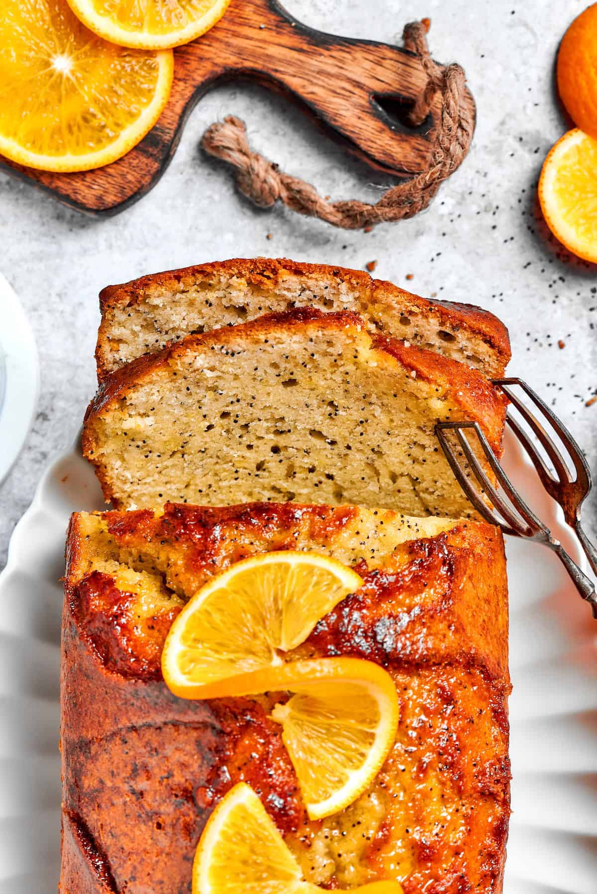 This moist orange poppy seed bread is soaked in a zippy citrus glaze for a bright, tender, bakery-style loaf perfect for brunch or gifting. Tongs picking up slice of orange glazed poppy bread from loaf.