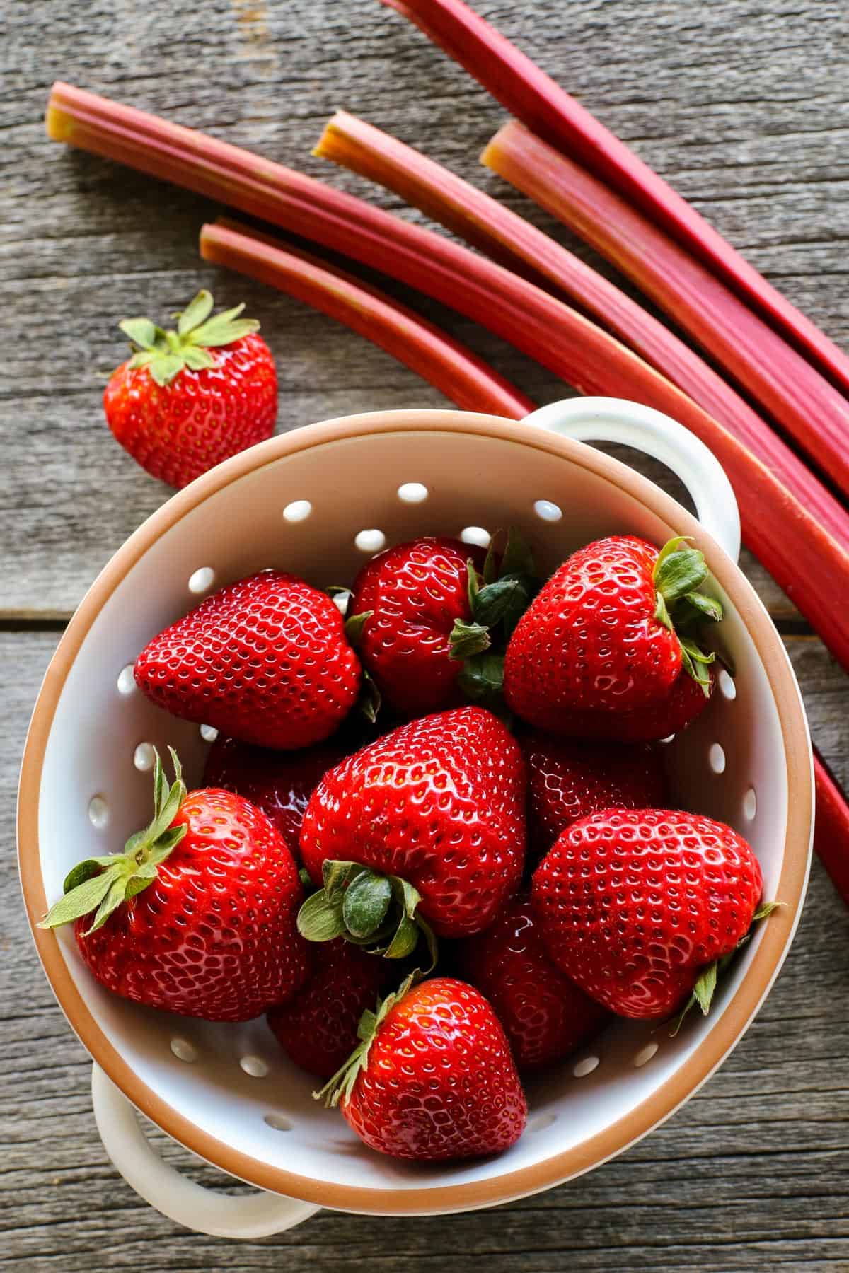 This Strawberry Rhubarb Crisp recipe features strawberries and rhubarb in a thick, jammy filling that's finished with a buttery oats topping. Strawberries are placed in in a colander alongside stalks of rhubarb.