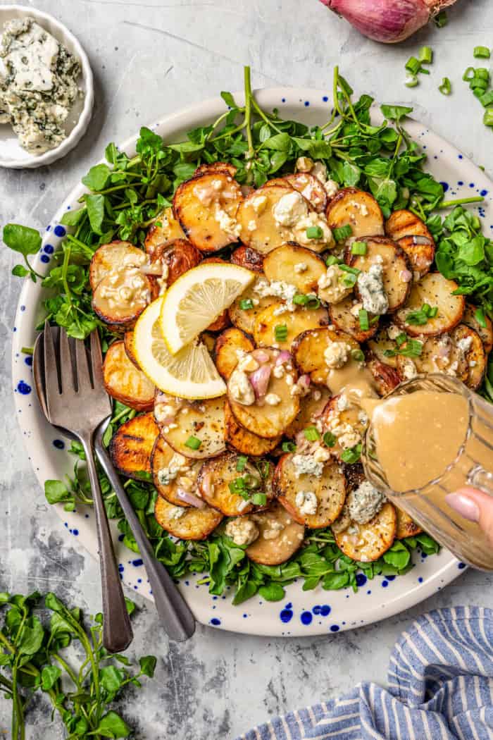 With smoky red potatoes and a rich blue cheese vinaigrette, this grilled potato salad recipe is so delicious, it might just steal the show! Pouring blue cheese vinaigrette onto platter of grilled potato salad