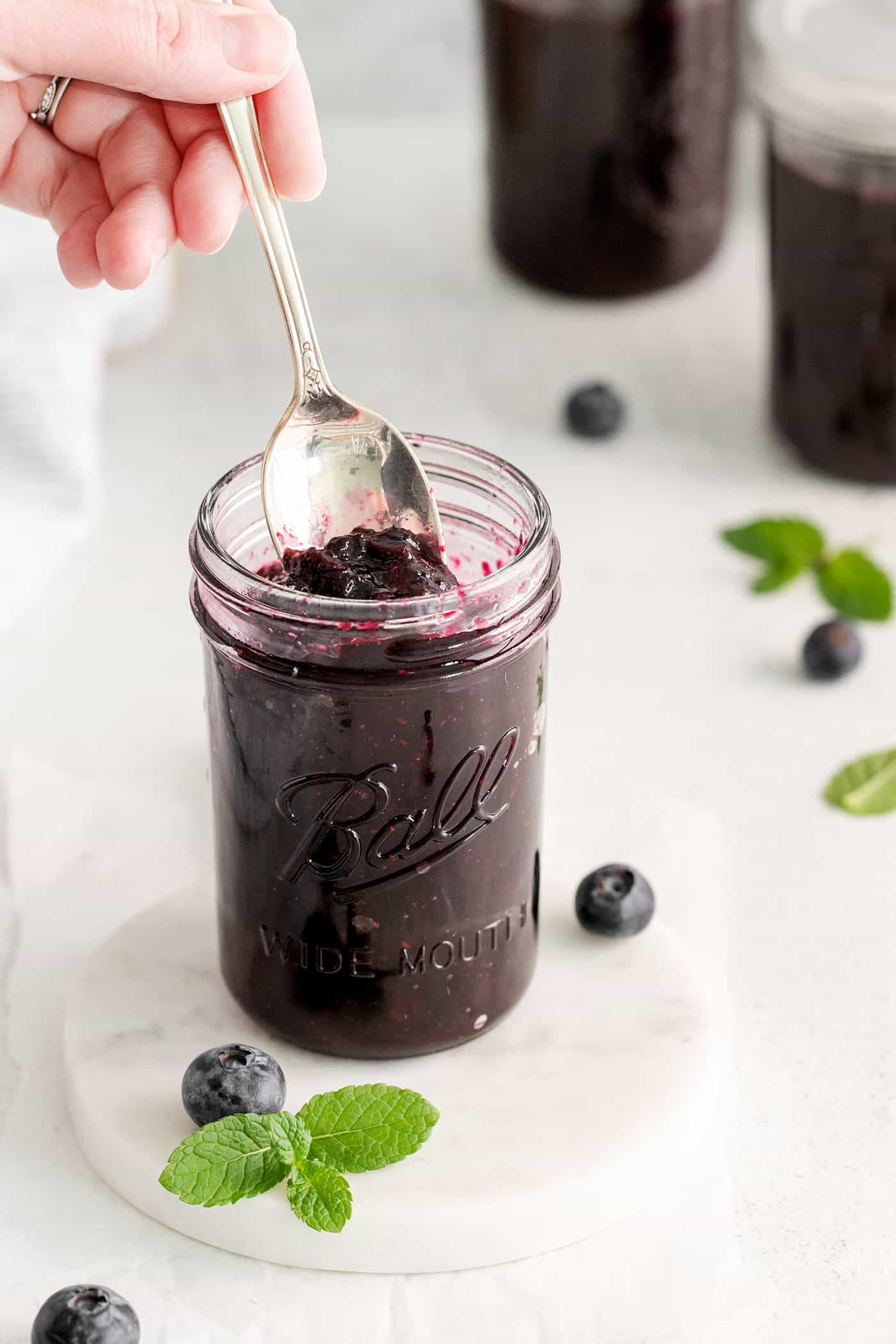 Made with just 4 ingredients, this Homemade Blueberry Jam with Mint is dark and fruity, with a sweet but cool and refreshing flavor. A hand holding a spoon in a jar of jam