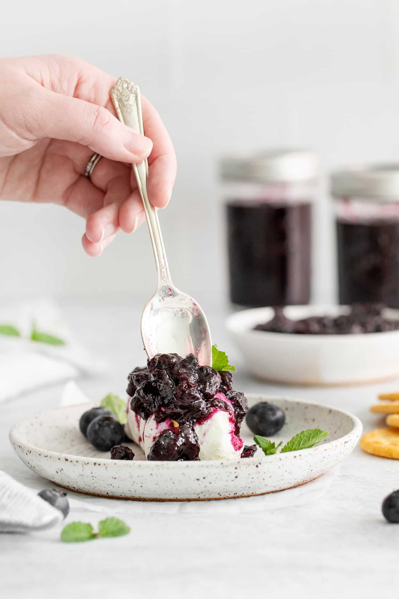 Made with just 4 ingredients, this Homemade Blueberry Jam with Mint is dark and fruity, with a sweet but cool and refreshing flavor. A hand spooning blueberry jam over ice cream