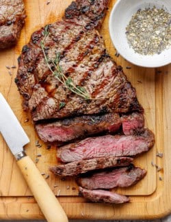 Sliced grilled ribeye steak on cutting board with knife and bowl of steak seasoning.