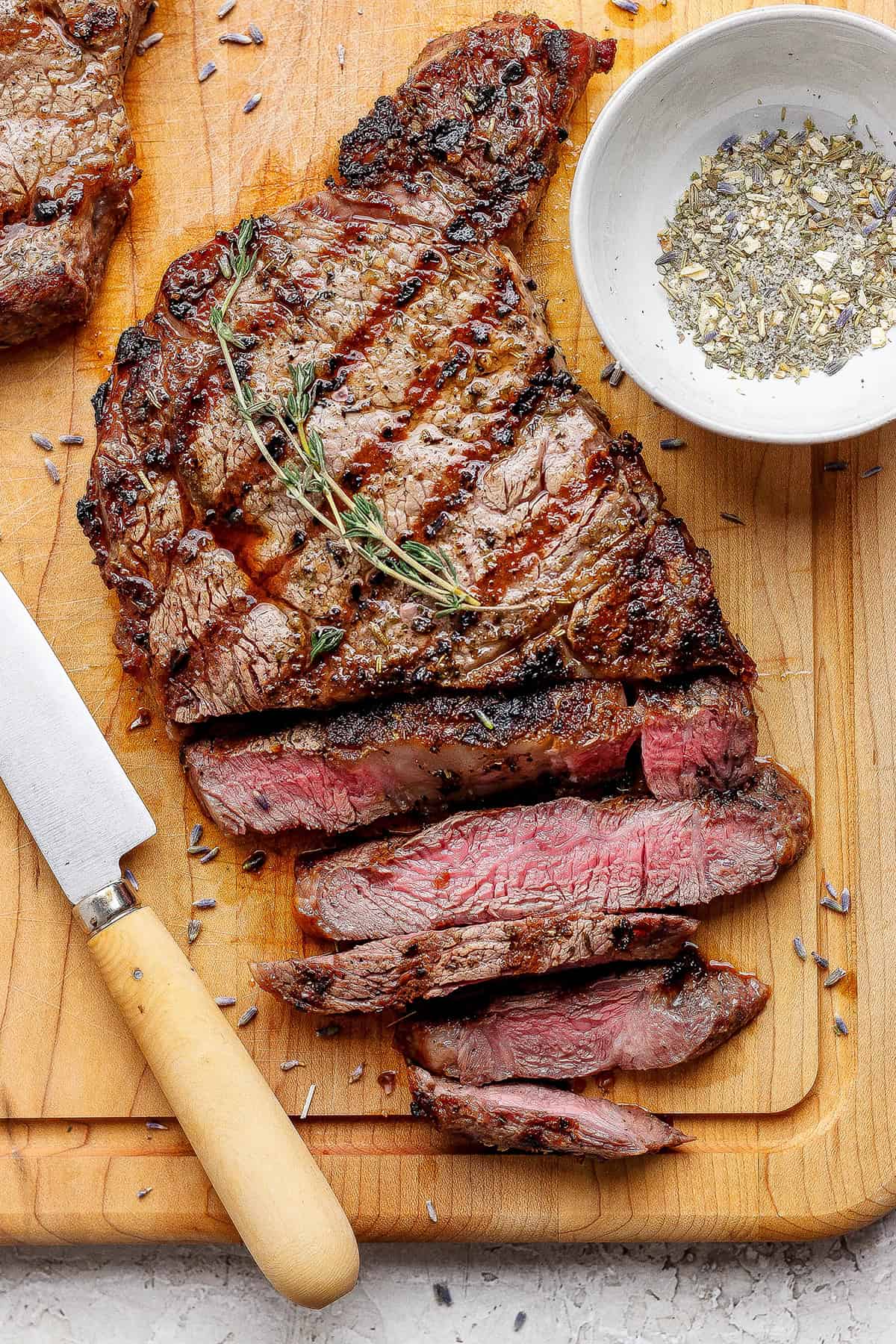 This Grilled Ribeye Recipe will show you the secret to perfectly grilled steak every time! It's tender, juicy, and packed with savory flavor. Sliced grilled ribeye steak on cutting board with knife and bowl of steak seasoning.