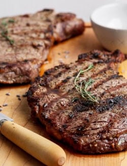 Two grilled ribeye steaks on cutting board with knife.