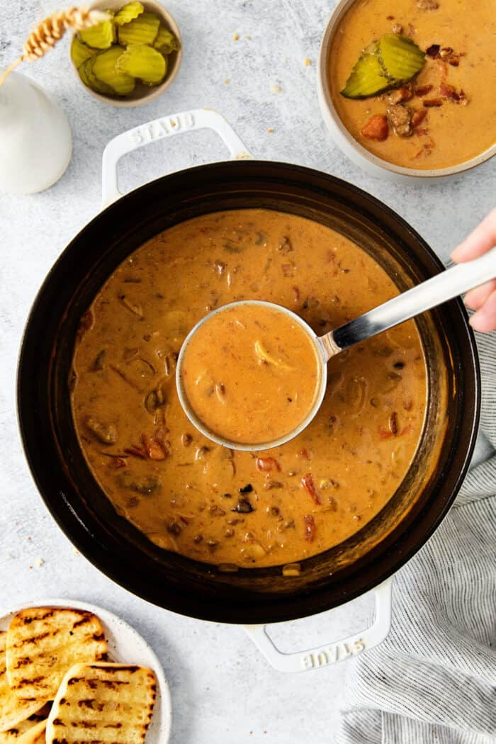 This Bacon Cheeseburger Soup is loaded with your favorite burger ingredients and toppings, like bacon, mushrooms, and a handful of seasonings. Top with shredded cheese and dill pickles for a soup that tastes just like your favorite cheeseburger! A ladle of soup over a pot of bacon cheeseburger soup