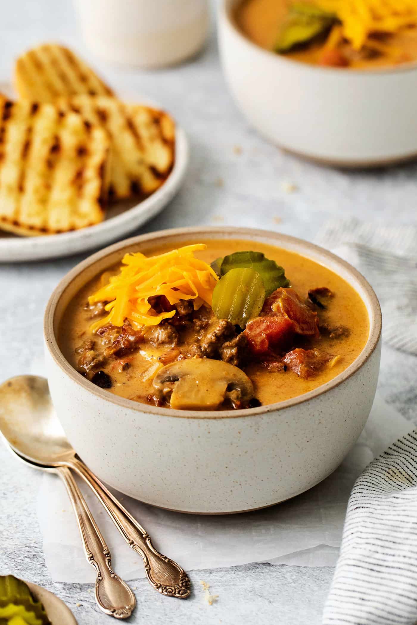 This Bacon Cheeseburger Soup is loaded with your favorite burger ingredients and toppings, like bacon, mushrooms, and a handful of seasonings. Top with shredded cheese and dill pickles for a soup that tastes just like your favorite cheeseburger! An angled view of a bowl of cheeseburger soup
