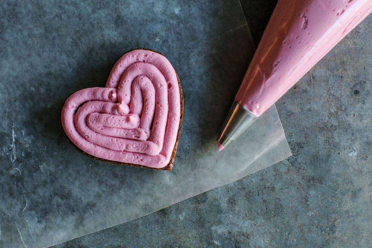 This heart shaped brownies recipe sandwiched with tangy raspberry-lemon buttercream is a super cute, chocolatey Valentine's Day treat. Piping raspberry frosting over a brownie heart.
