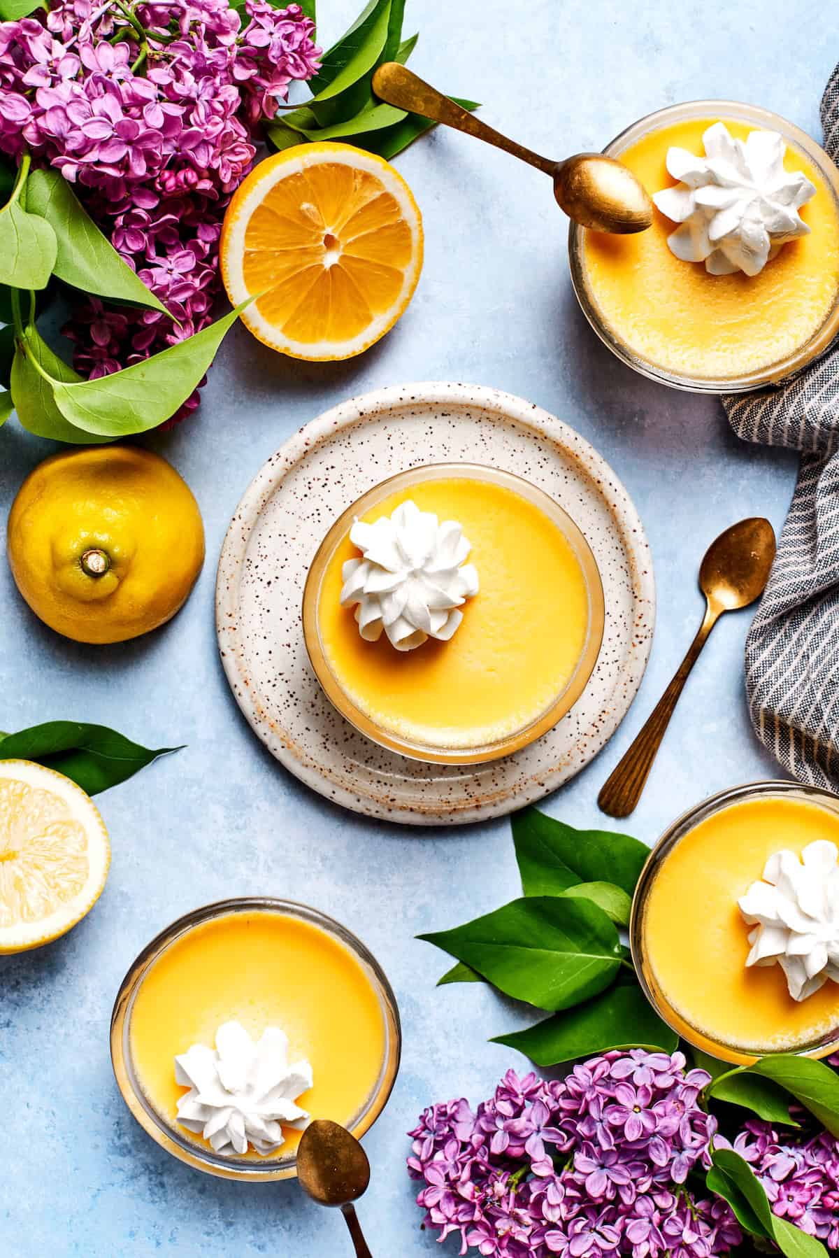 Overhead view of 4 lemon custard cups with whipped cream, surrounded by fresh lemons and lilacs