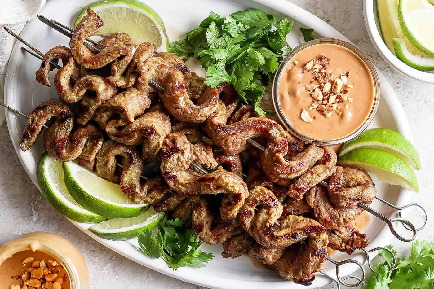 Tender beef is marinated in coconut milk, threaded onto skewers, and grilled to perfection in this coconut beef satay recipe. So flavorful! Overhead view of coconut beef satay on platter with bowl of peanut sauce, cilantro, and lime wedges