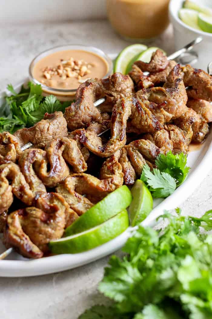 Tender beef is marinated in coconut milk, threaded onto skewers, and grilled to perfection in this coconut beef satay recipe. So flavorful! Coconut beef satay skewers on platter with peanut sauce, cilantro, and lime