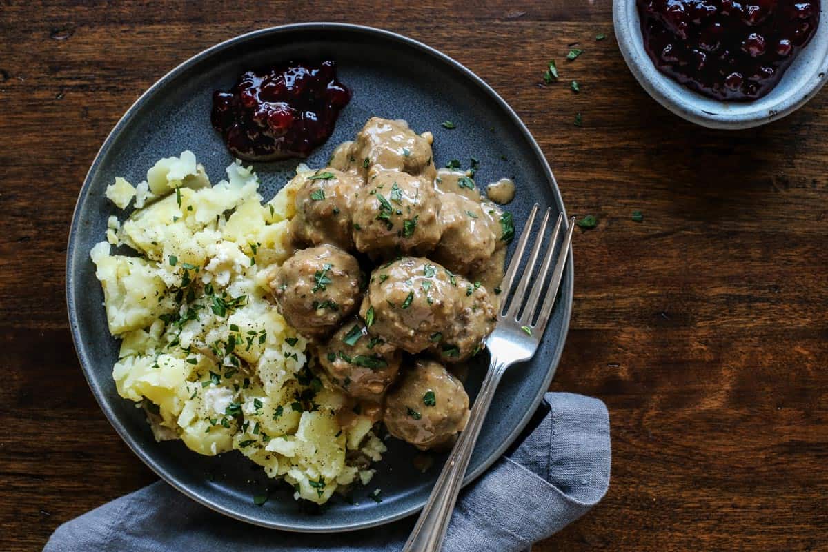 This easy Swedish meatballs recipe makes juicy, tender meatballs with a rich, creamy gravy. A homemade version of IKEA Swedish meatballs! Overhead view of Swedish meatballs and gravy served with potatoes and lingonberry jam on a stoneware plate with a fork.