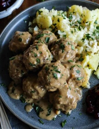 Swedish meatballs and gravy served with potatoes and lingonberry jam on a stoneware plate.
