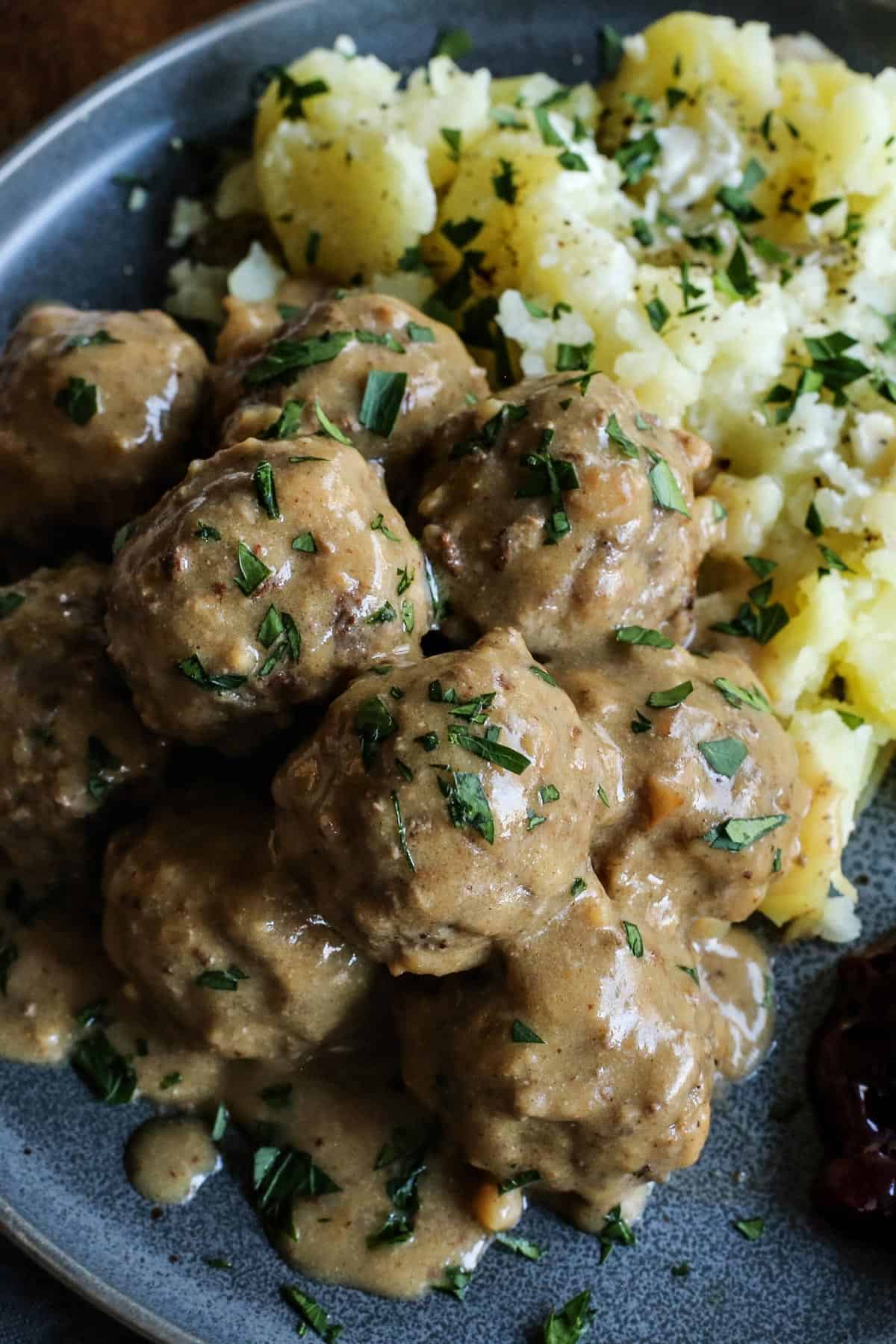 This easy Swedish meatballs recipe makes juicy, tender meatballs with a rich, creamy gravy. A homemade version of IKEA Swedish meatballs! Close up of homemade IKEA Swedish meatballs and gravy served with potatoes and lingonberry jam on a stoneware plate.