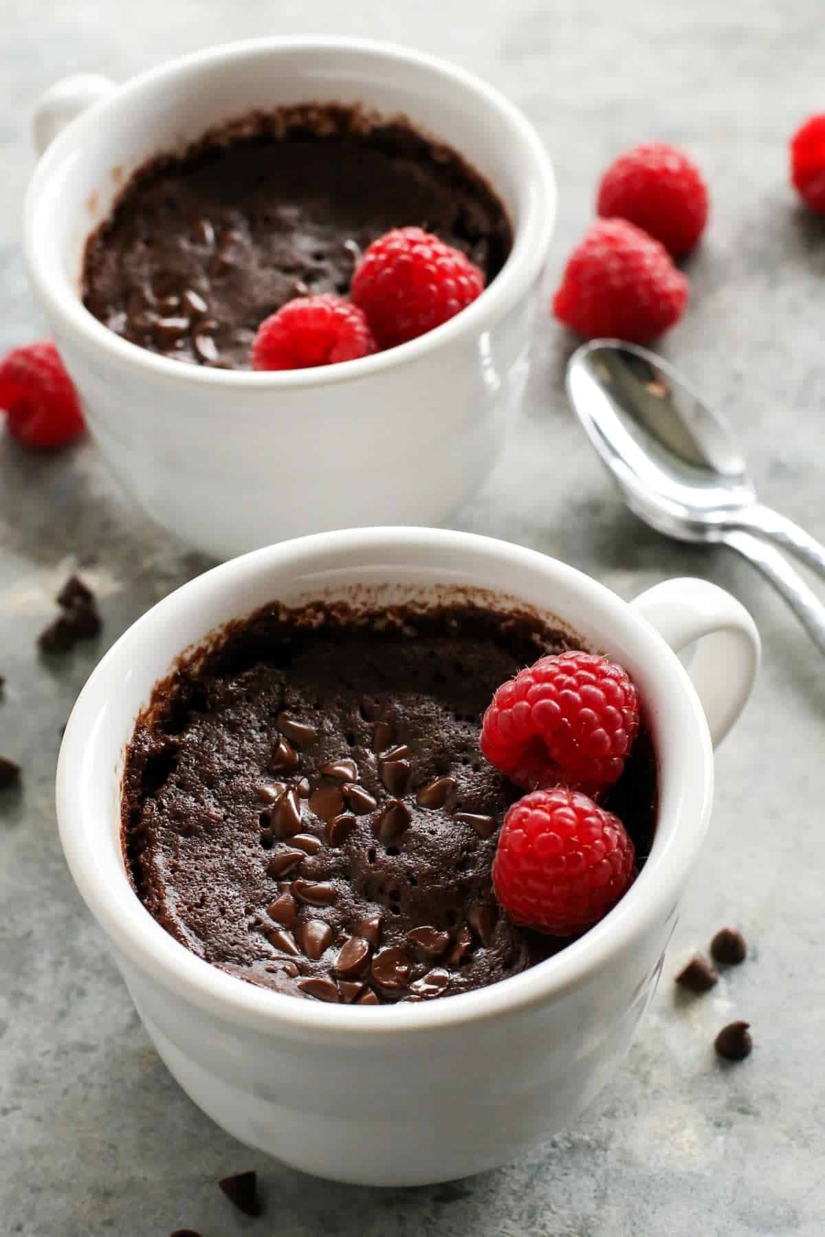 This gooey chocolate mug cake is fudgy, fluffy, and ready in minutes! Make this moist chocolate cake recipe with or without eggs. two white mugs of chocolate cake with fresh raspberries