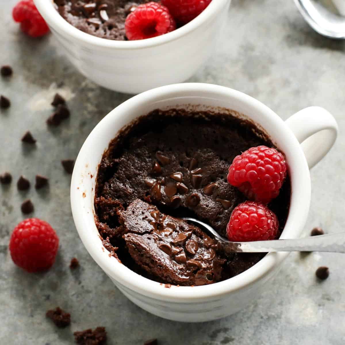 This gooey chocolate mug cake is fudgy, fluffy, and ready in minutes! Make this moist chocolate cake recipe with or without eggs. a spoon dipping into a white mug of chocolate cake with fresh raspberries