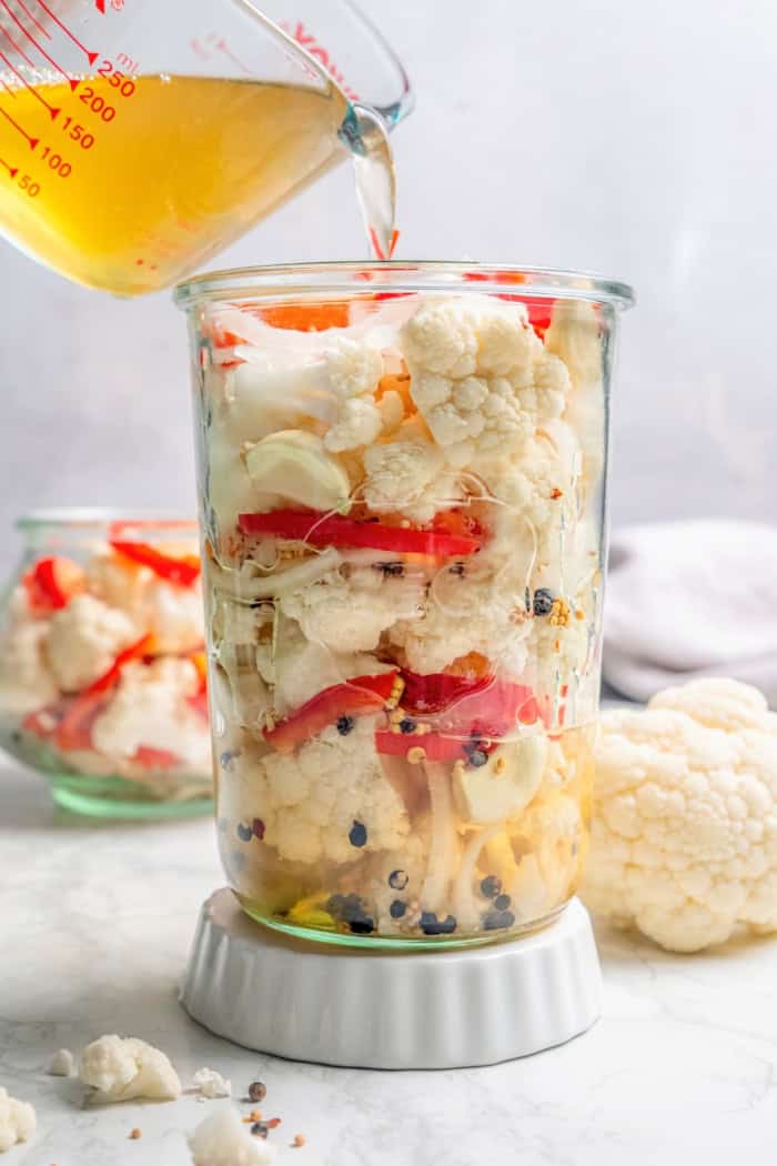 Pouring brine from liquid measuring cup into jar of cauliflower, onions, and red peppers