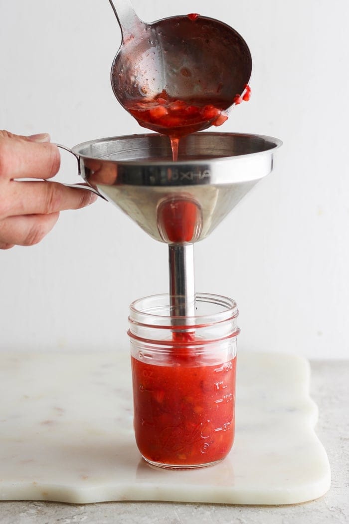 With the right balance of sweet and tart, this strawberry rhubarb freezer jam is a delight — and you don't need canning equipment to make it! Pouring strawberry rhubarb jam into jar with funnel