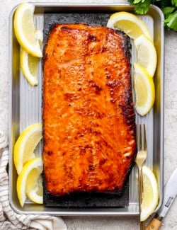 Overhead view of sweet and smoky grilled salmon set on cedar plank inside sheet pan with lemon wedges