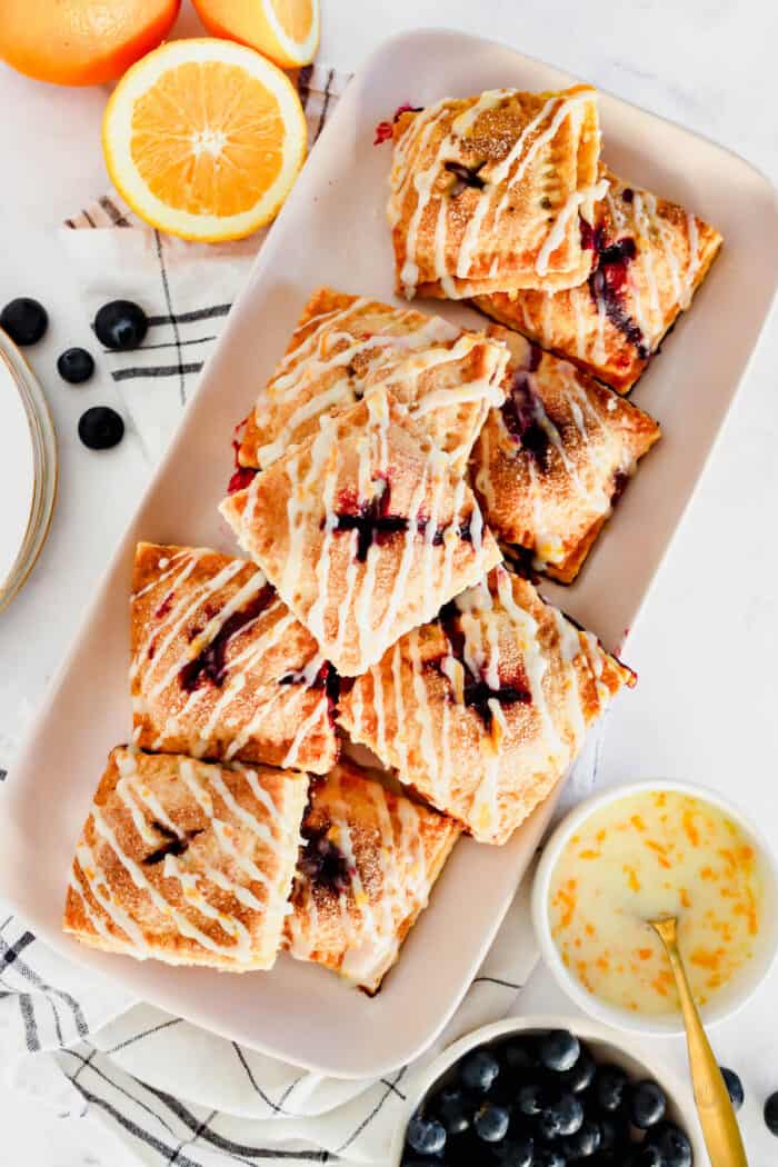 These cute blueberry hand pies combine a flaky pastry crust with sweet blueberry pie filling. It's so much easier than baking a whole pie! Overhead view of a platter of blueberry hand pies drizzled with orange glaze, next to small bowls with pie ingredients.