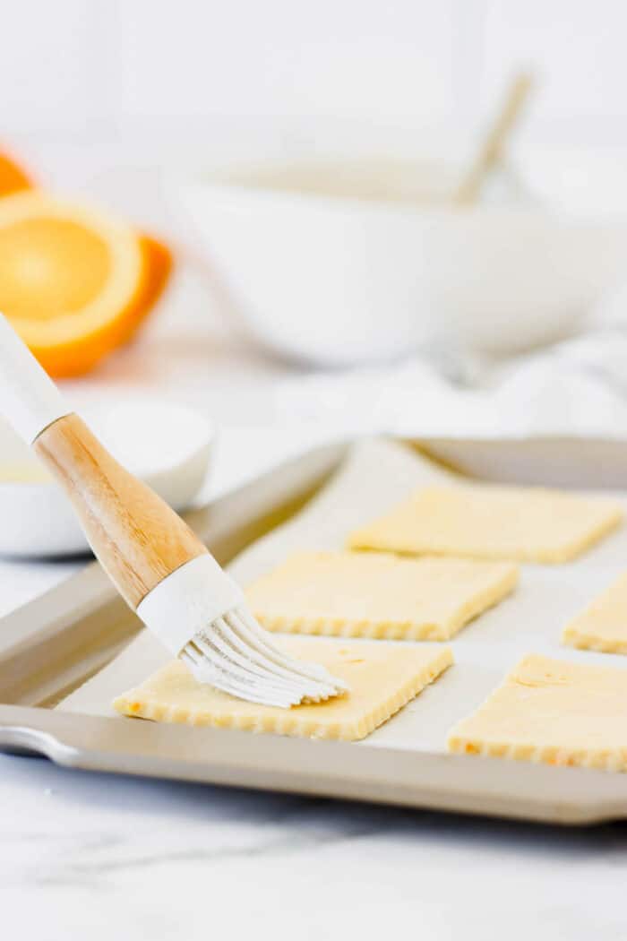 These cute blueberry hand pies combine a flaky pastry crust with sweet blueberry pie filling. It's so much easier than baking a whole pie! A basting brush brushing egg wash over squares of cut out pie dough on a lined baking sheet.