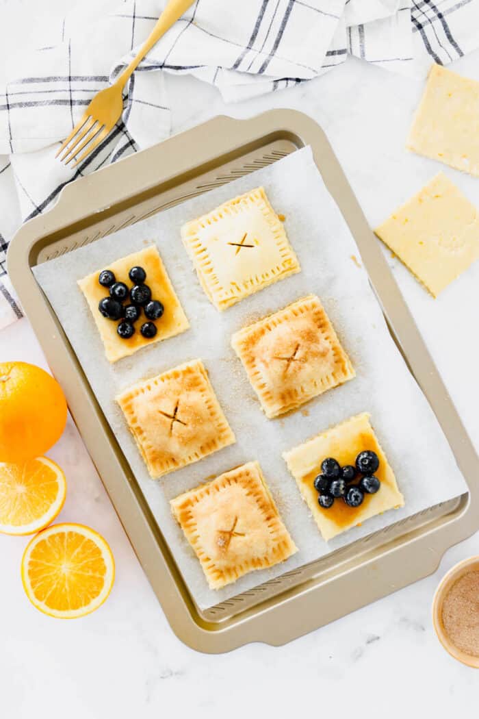 These cute blueberry hand pies combine a flaky pastry crust with sweet blueberry pie filling. It's so much easier than baking a whole pie! Overhead view of partially assembled blueberry hand pies on a lined baking sheet.