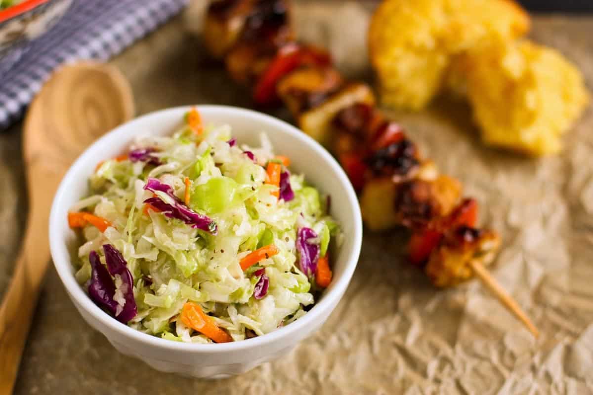 This Sweet Onion Vinegar Coleslaw recipe is fresh, bright & tangy. No mayo here! It has a coleslaw dressing that's seriously good! coleslaw salad in a small white bowl, chicken kebab, and cornbread muffin