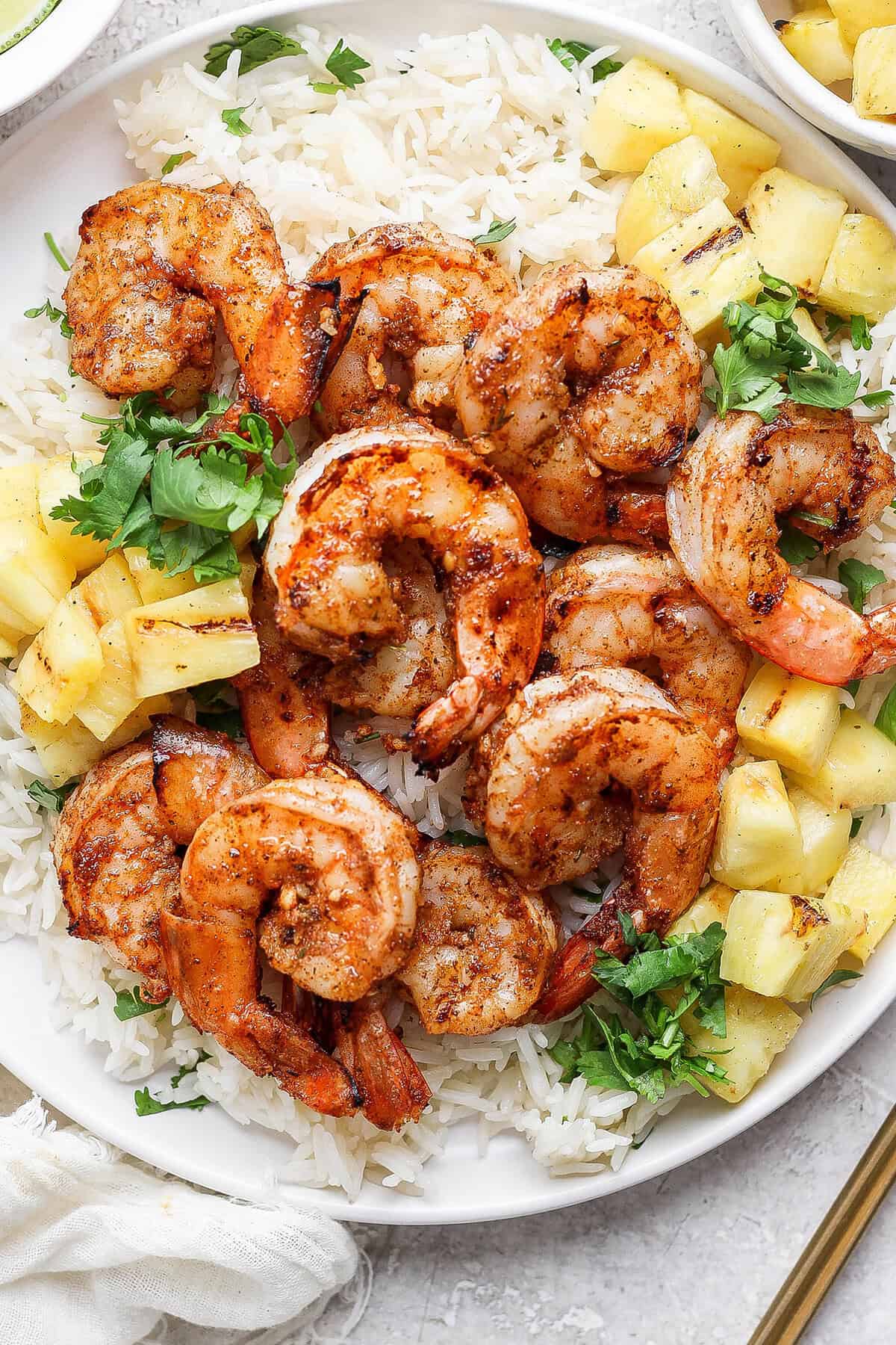 Enjoy this grilled jerk shrimp recipe as a tantalizing appetizer, gorgeous salad topper, or main entrée. It's quick, easy, and so flavorful! Overhead view of grilled jerk shrimp on plate with cilantro lime rice and pineapple