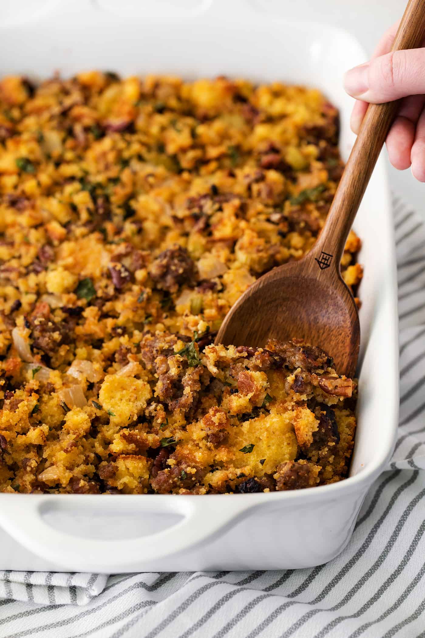 A wooden spoon serving cornbread sausage dressing from a white casserole dish.