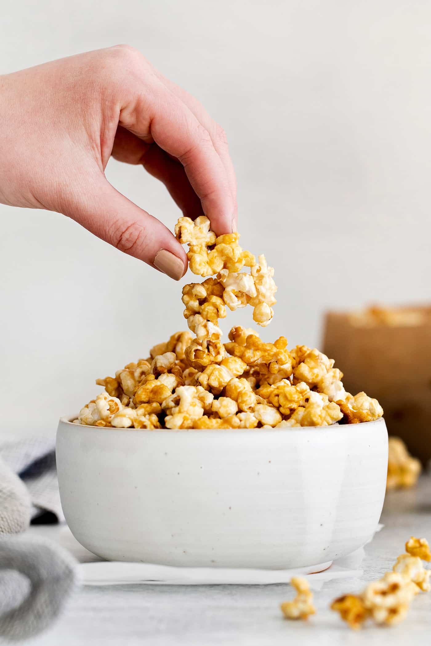 Microwave Caramel Popcorn is ready in just 15 minutes, with a handful of ingredients, a microwave, and a brown paper bag. A hand grabbing a piece of caramel popcorn from a white bowl