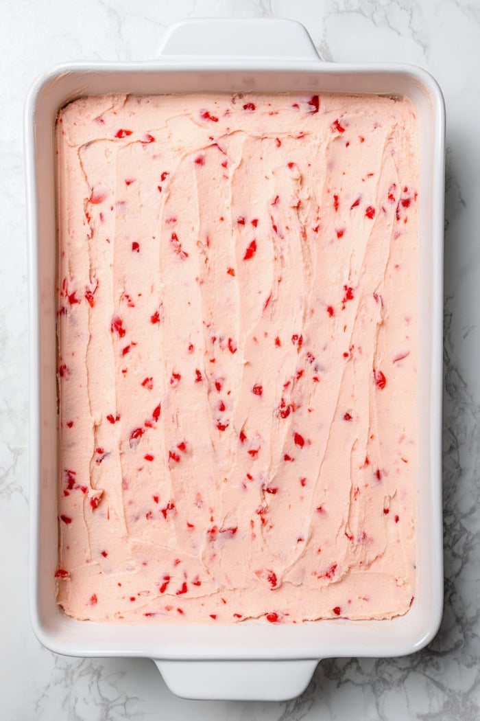 These fudgy cherry brownies are layered with fluffy cherry buttercream and smooth chocolate glaze for an indulgent bakery-style dessert! Cherry buttercream spread onto pan of brownies.