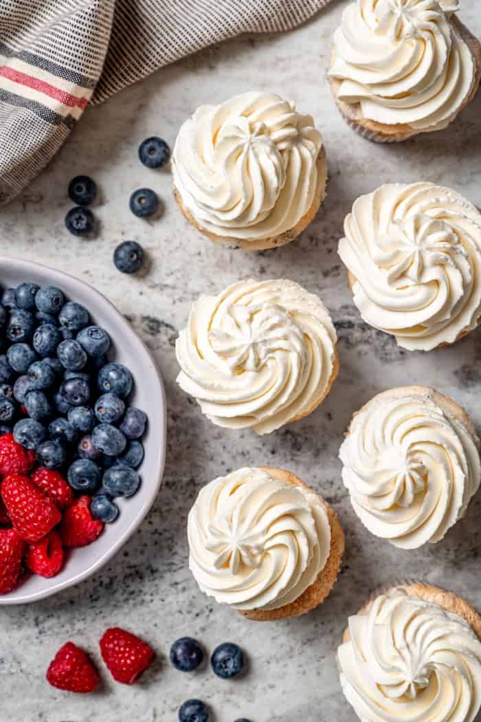 These Angel Food Cupcakes are dotted with mini chocolate chips and topped with vanilla cream and fresh berries. A light, summery treat! Overhead view of frosted angel food cupcakes with bowl of fresh berries