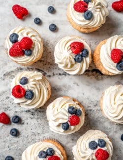 Overhead view of angel food cupcakes topped with frosting and fresh berries on marble countertop