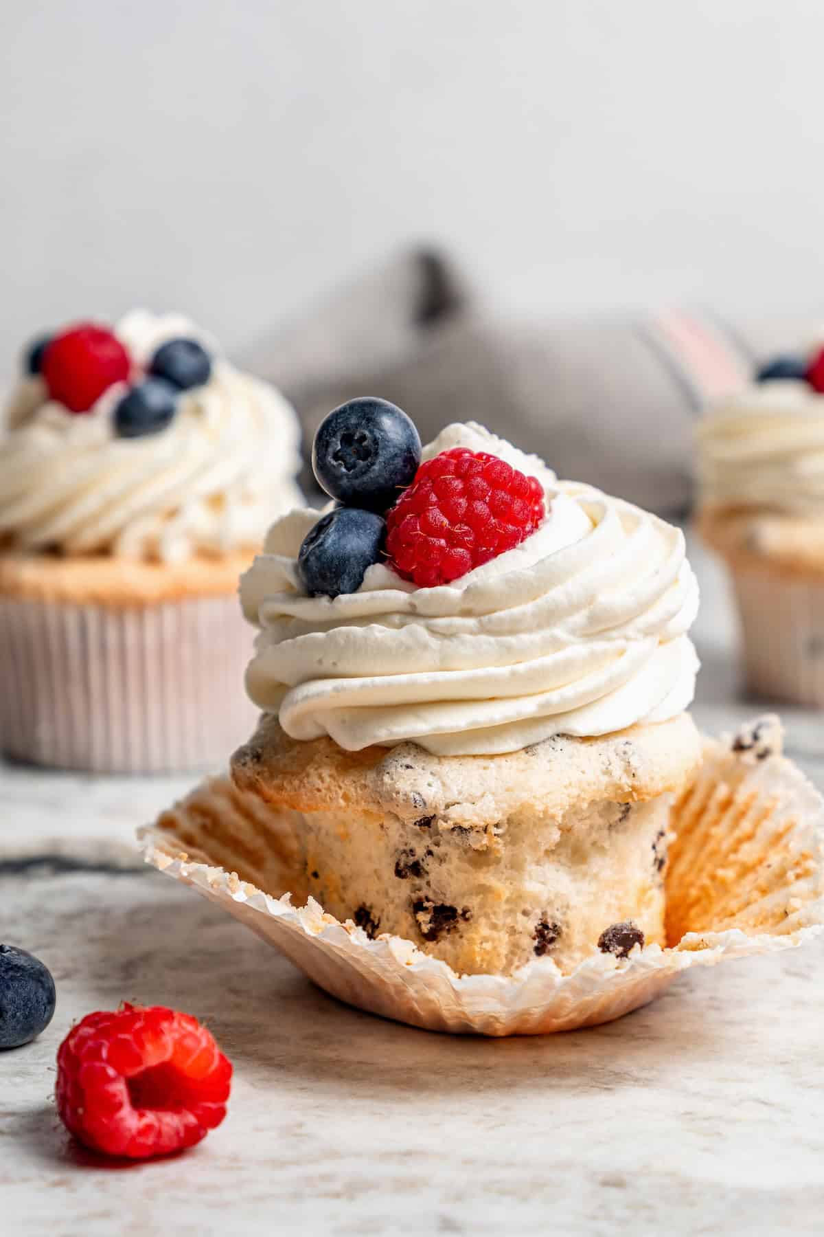 These Angel Food Cupcakes are dotted with mini chocolate chips and topped with vanilla cream and fresh berries. A light, summery treat! Chocolate chip angel food cupcake with paper wrapper pulled off