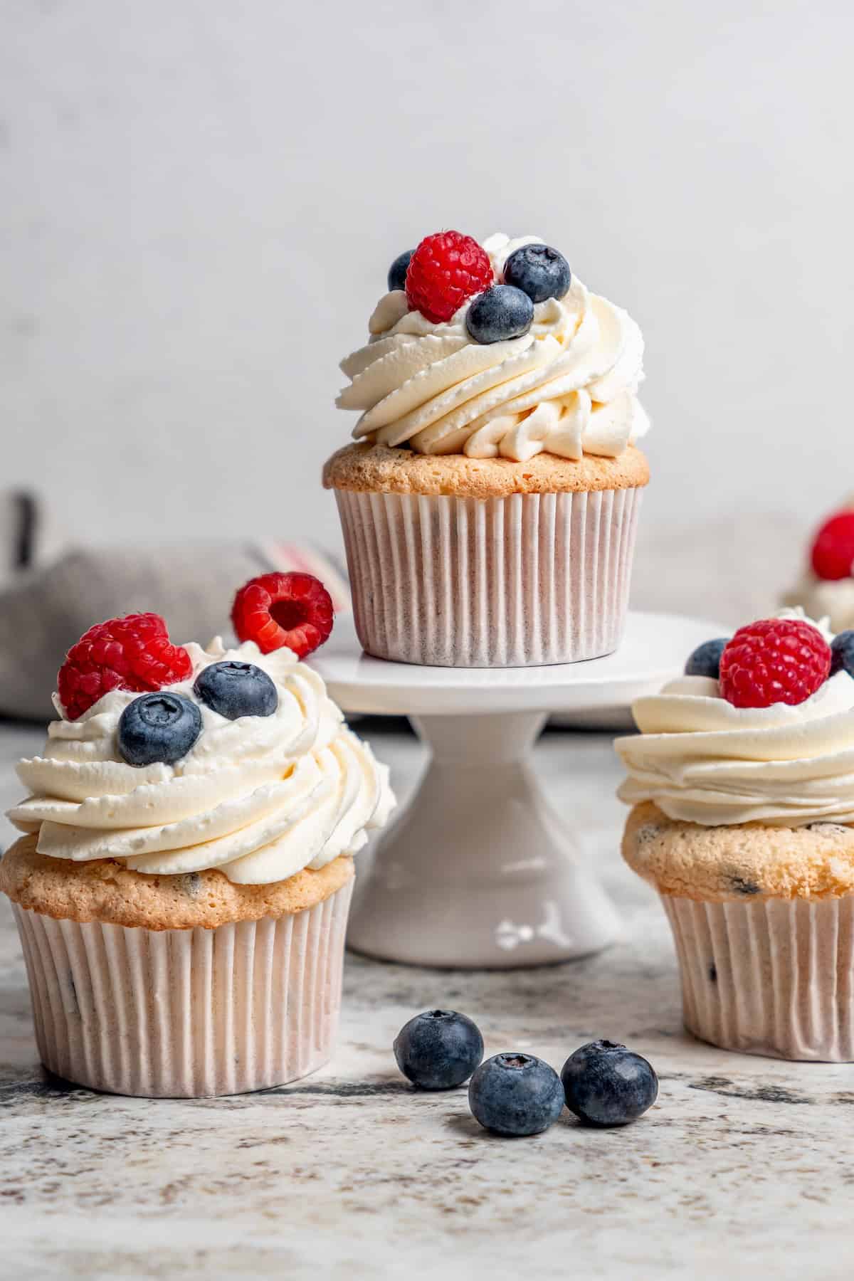 These Angel Food Cupcakes are dotted with mini chocolate chips and topped with vanilla cream and fresh berries. A light, summery treat! Angel food cupcake on stand, with other cupcakes on counter