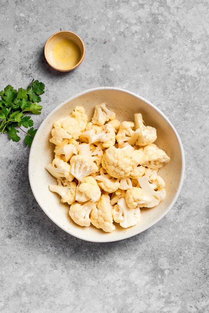 This easy roasted cauliflower is jazzed up with pepitas, cumin, smoky ancho, garlic, and lime. Crispy on the outside, tender inside! Bowl of raw cauliflower florets and bowl of oil.