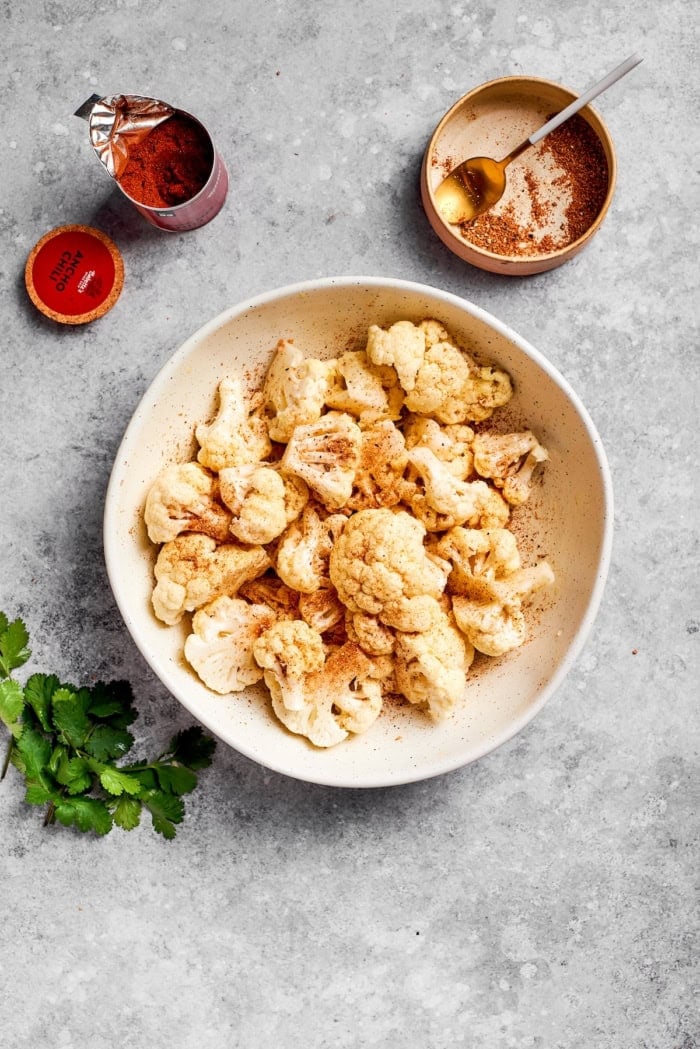 This easy roasted cauliflower is jazzed up with pepitas, cumin, smoky ancho, garlic, and lime. Crispy on the outside, tender inside! Bowl of seasoned raw cauliflower.